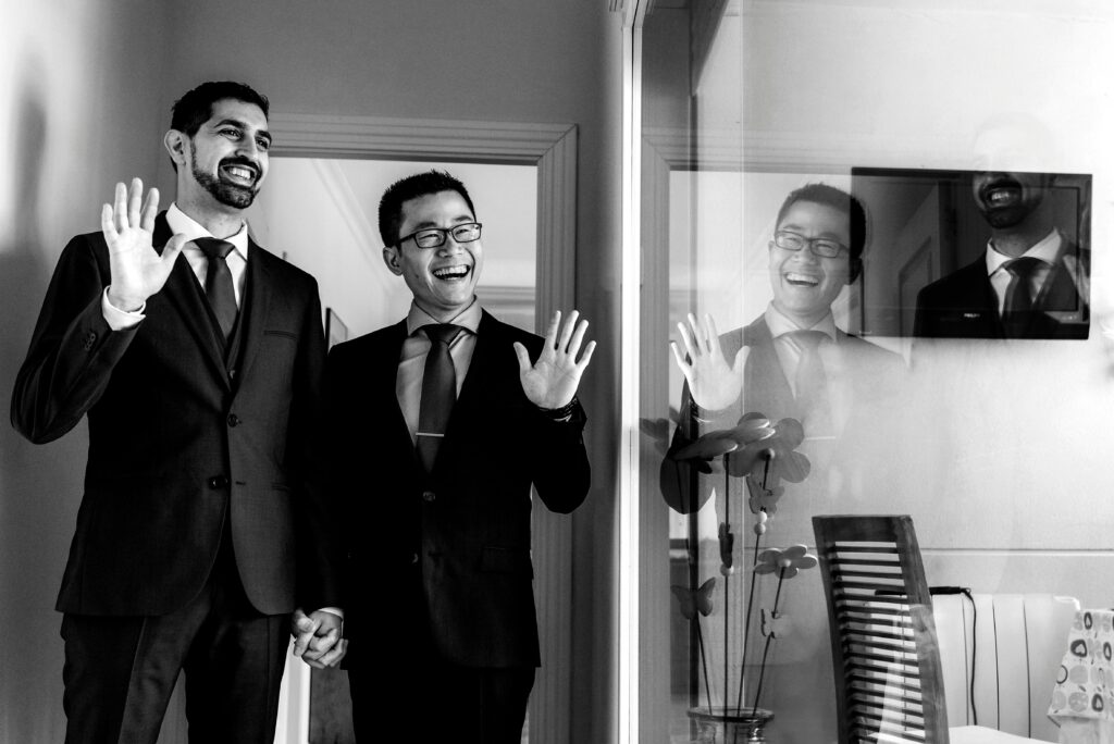 A joyful same-sex couple in suits waving, captured indoors in a black and white photo.